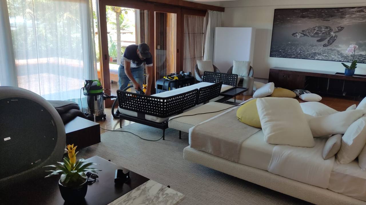 Profissional da Mac Clean, de uniforme e calça, higienizando um grande conjunto de estofados claros em sala residencial ampla de alto padrão, com o equipamento profissional ao lado e um quadro de tartaruga ao fundo