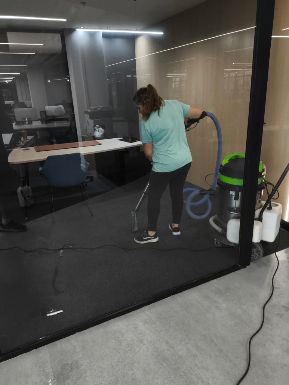 Profissional Mac Clean executando a extração em carpete de escritório, vista pela divisória de vidro, com a máquina extratora à direita